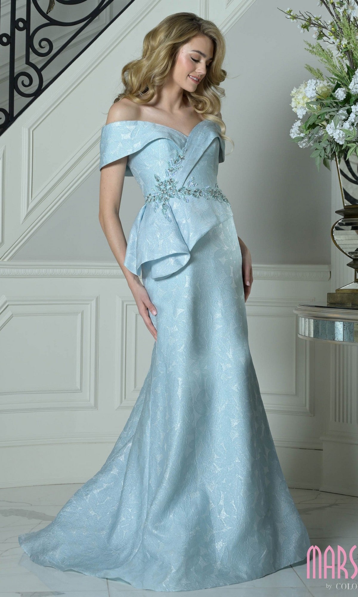 A woman in an aqua Marsoni MV1397 long formal dress with floral embellishments and an asymmetrical peplum waist stands indoors near a staircase and a table adorned with flowers.